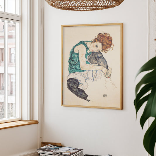 Egon Schiele zittende vrouw poster aan witte muur, met links een raam en rieten lampenkap erboven – plant rechts in beeld