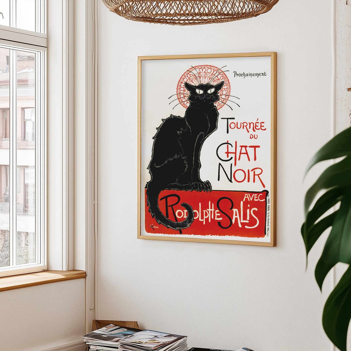 Zwarte kat poster aan witte muur, met links een raam en rieten lampenkap erboven – plant rechts in beeld.