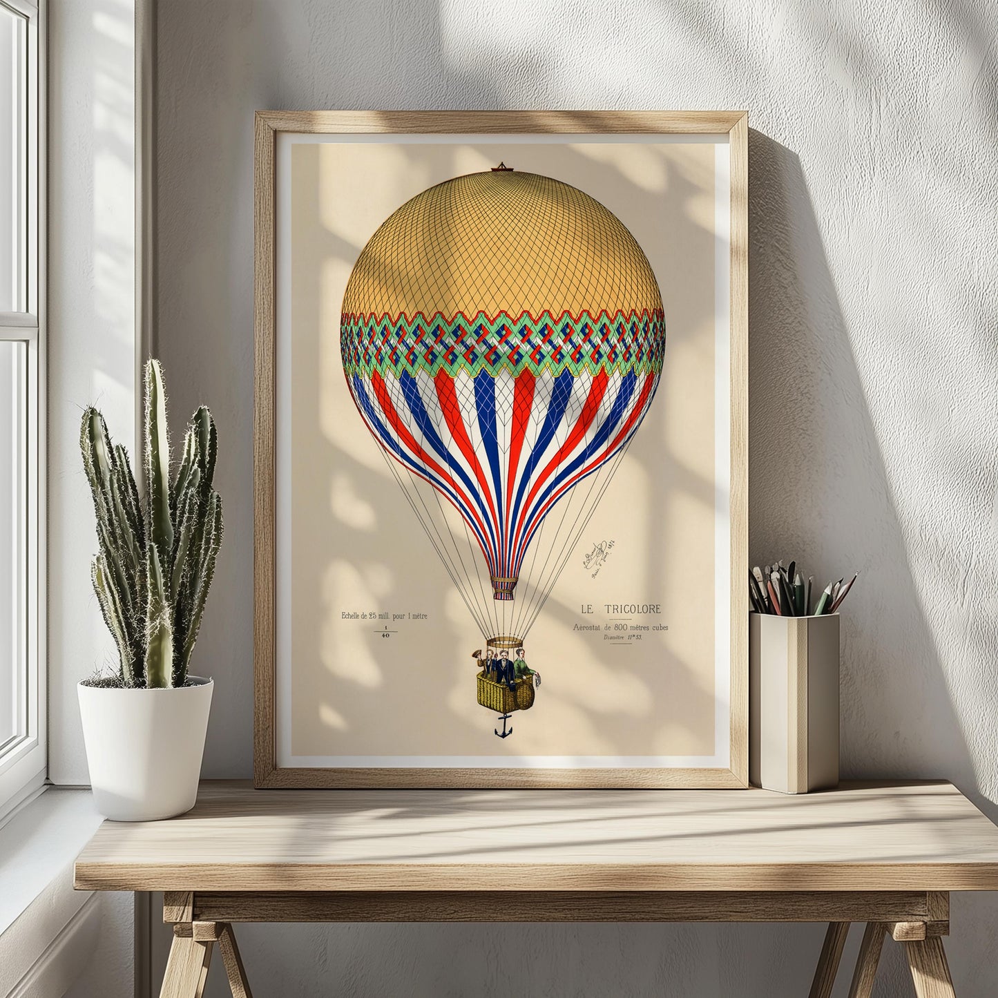 Ingelijste luchtballonposter op houten tafeltje, met links een cactus en rechts een pennenbakje – warme, natuurlijke sfeer.